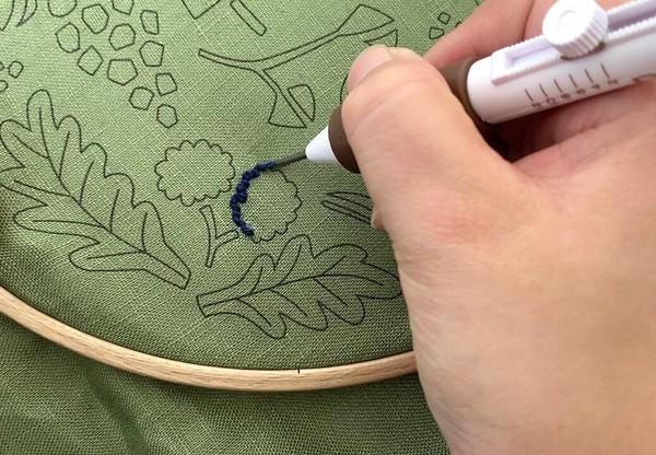 A hand is using a pen to draw on a green fabric with a black outline, creating a pattern or design.