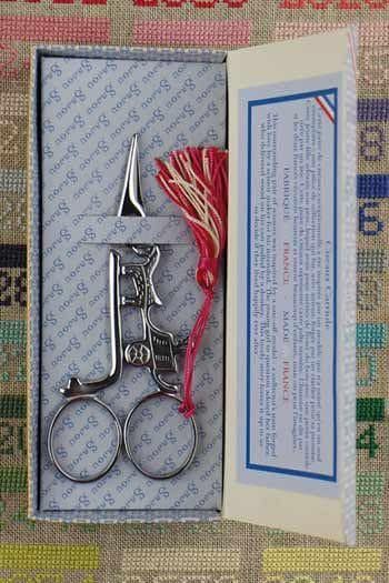 A pair of silver scissors with a red tassel, placed in a box with a blue and white label.