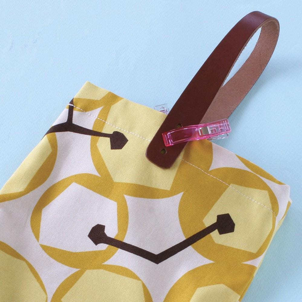 A yellow and white patterned fabric with a brown leather handle attached to it, along with a pink plastic clip.