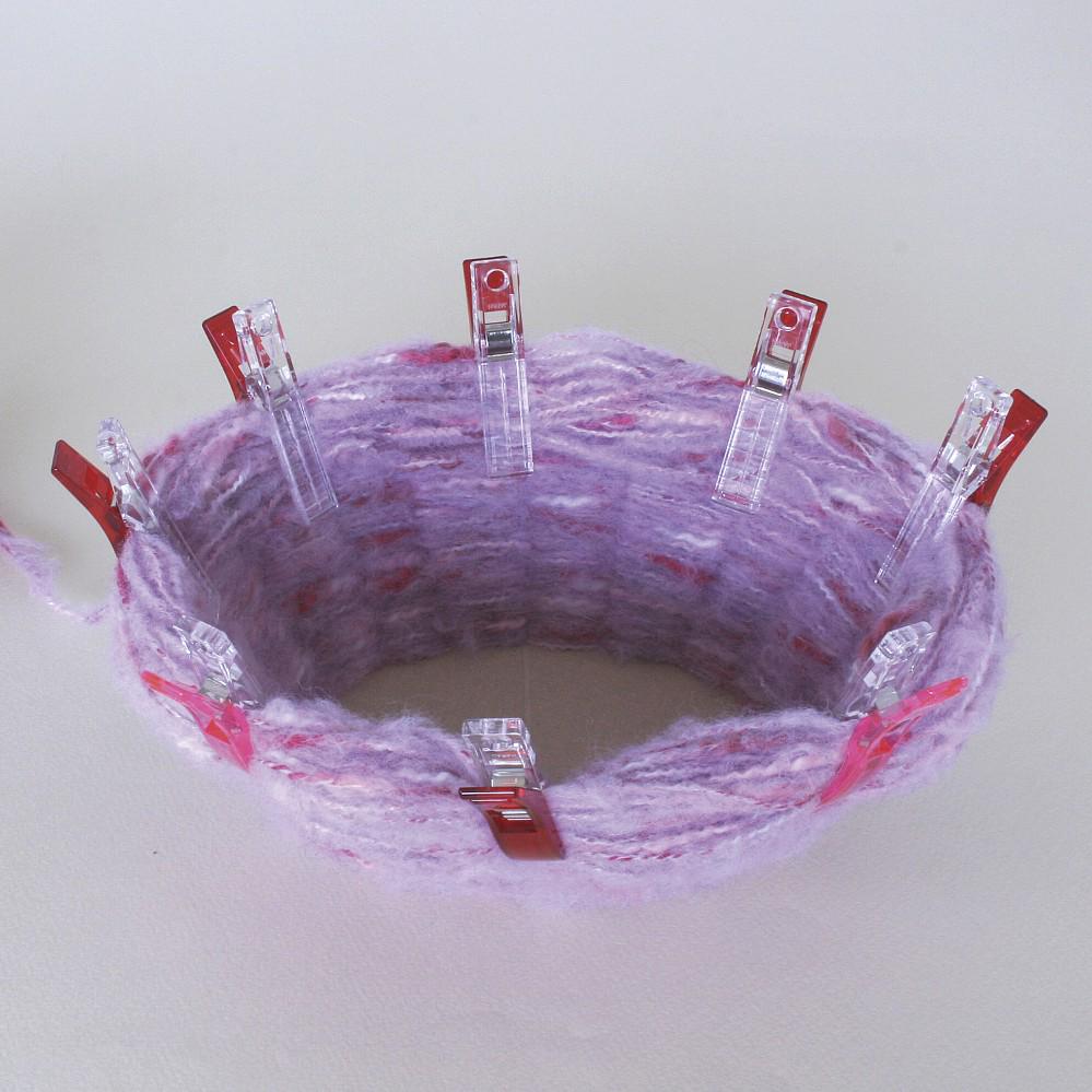 a close-up view of a purple, knitted or crocheted item with red plastic clips attached to it, likely for securing or attaching the yarn.