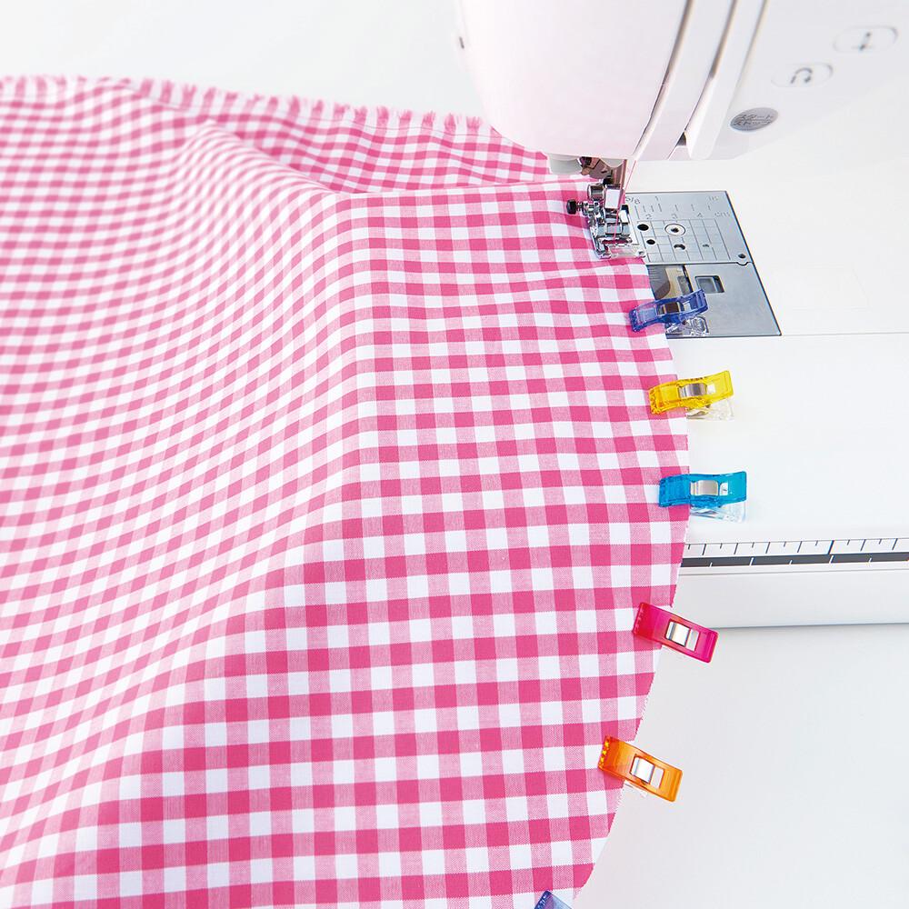 A pink and white checkered fabric is being sewn by a sewing machine, with colorful plastic clips attached to the edges of the fabric.