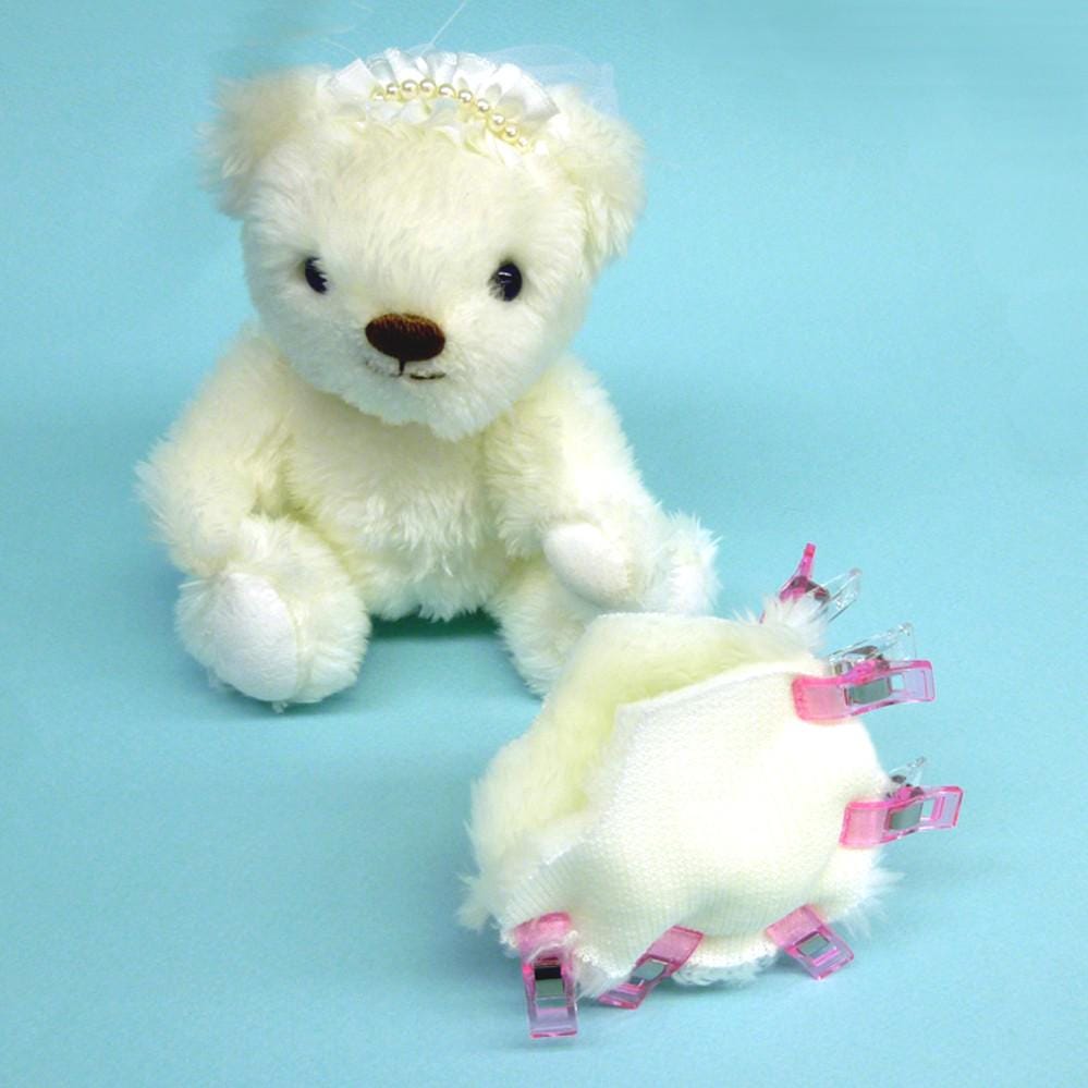 A white teddy bear wearing a white veil sits next to a white fabric item with pink clips attached to it, against a light blue background.