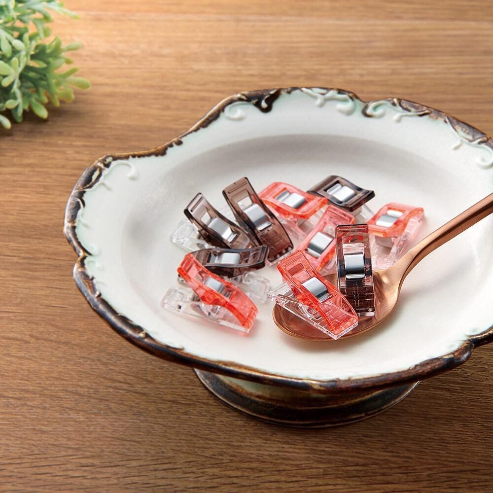 A white bowl with a scalloped edge is filled with red and orange sewing clips, and a spoon is resting inside the bowl.