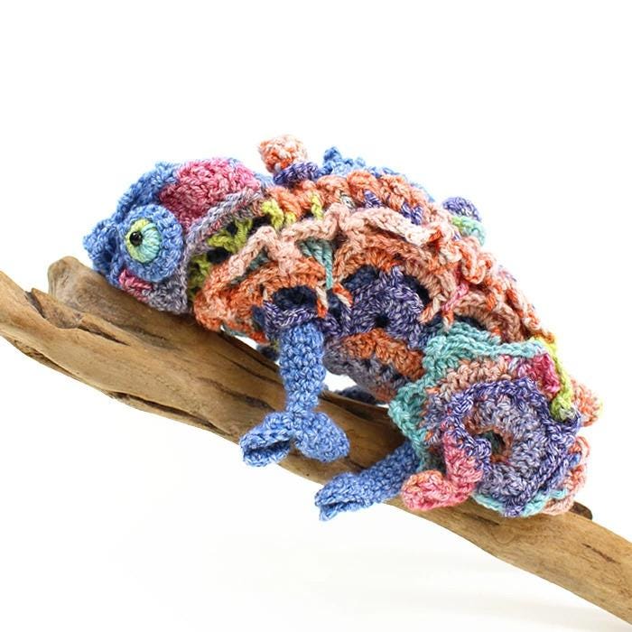 A crocheted chameleon toy is perched on a wooden branch.