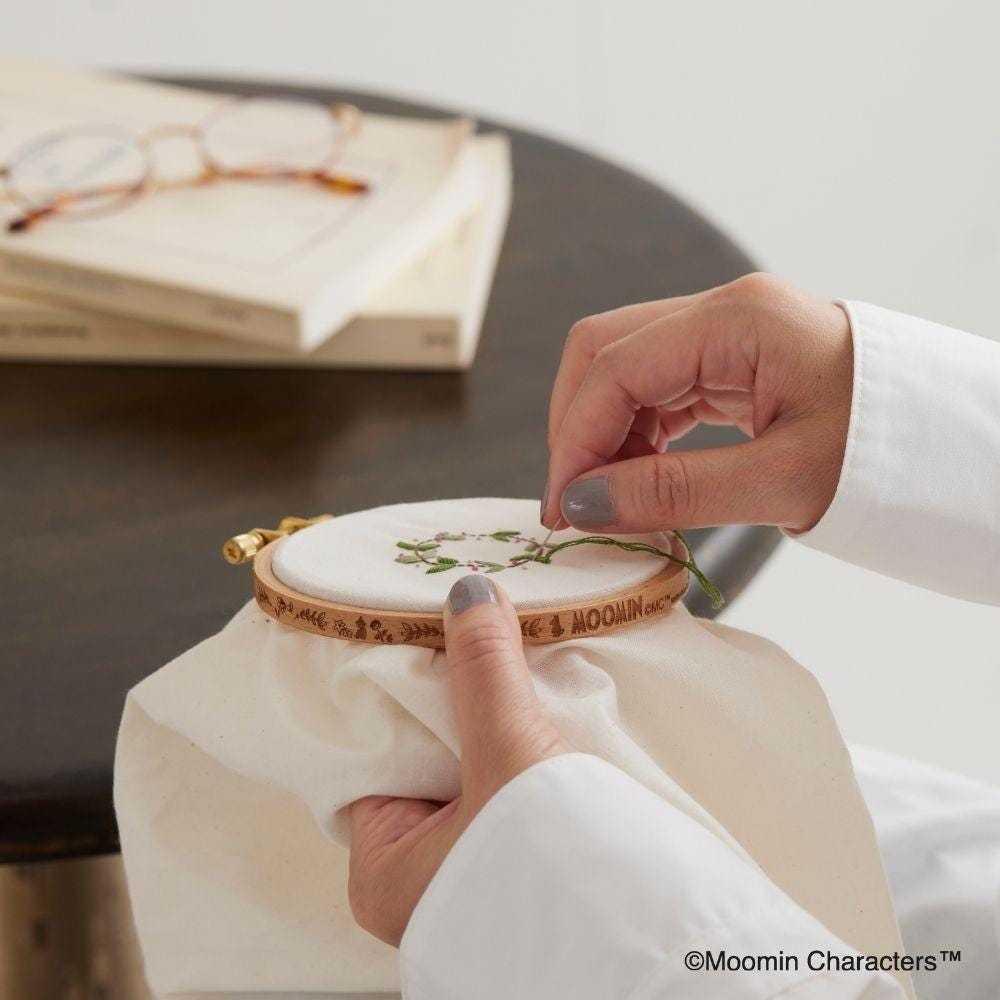 A person's hands are carefully threading a needle through a small embroidery hoop, which is adorned with a delicate floral design.