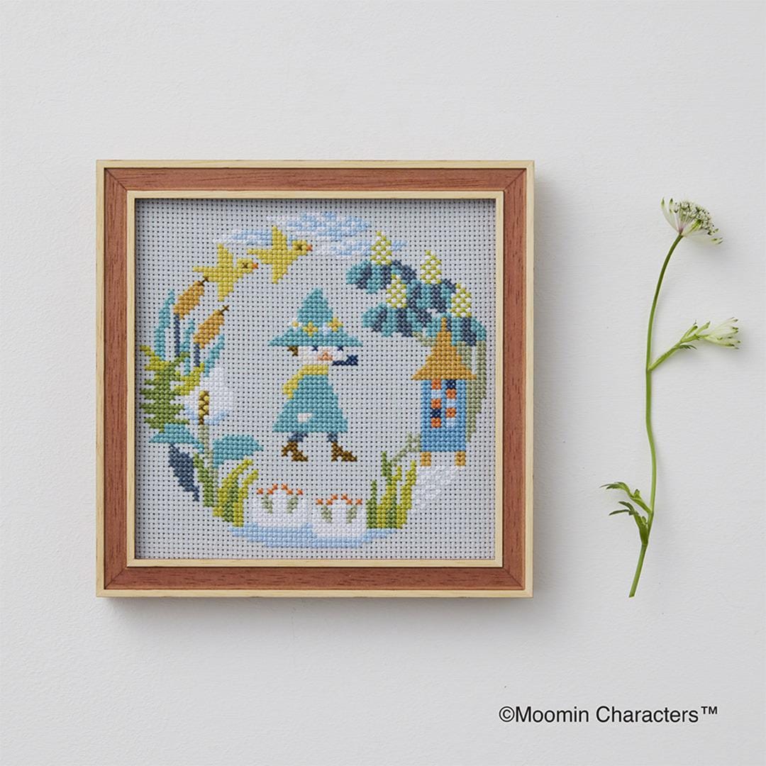 a cross-stitch design of a whimsical scene with a character, a bird, and a flower, all set against a light blue background. The design is framed and displayed on a white wall, accompanied by a small plant on the right side.