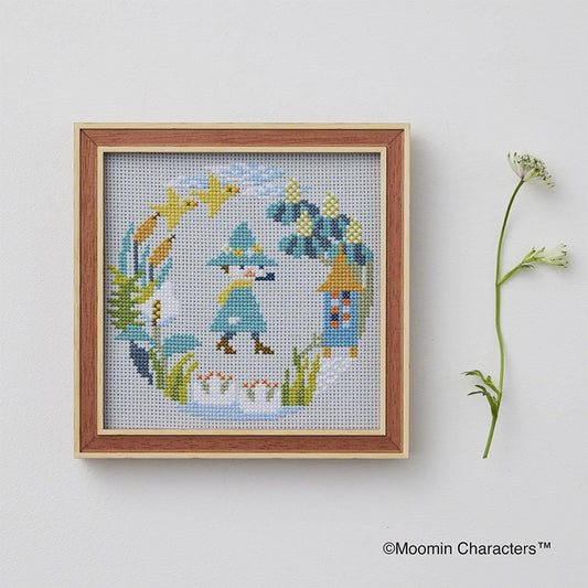 a cross-stitch design of a whimsical scene with a character, a bird, and a flower, all set against a light blue background. The design is framed and displayed on a white wall, accompanied by a small plant on the right side.