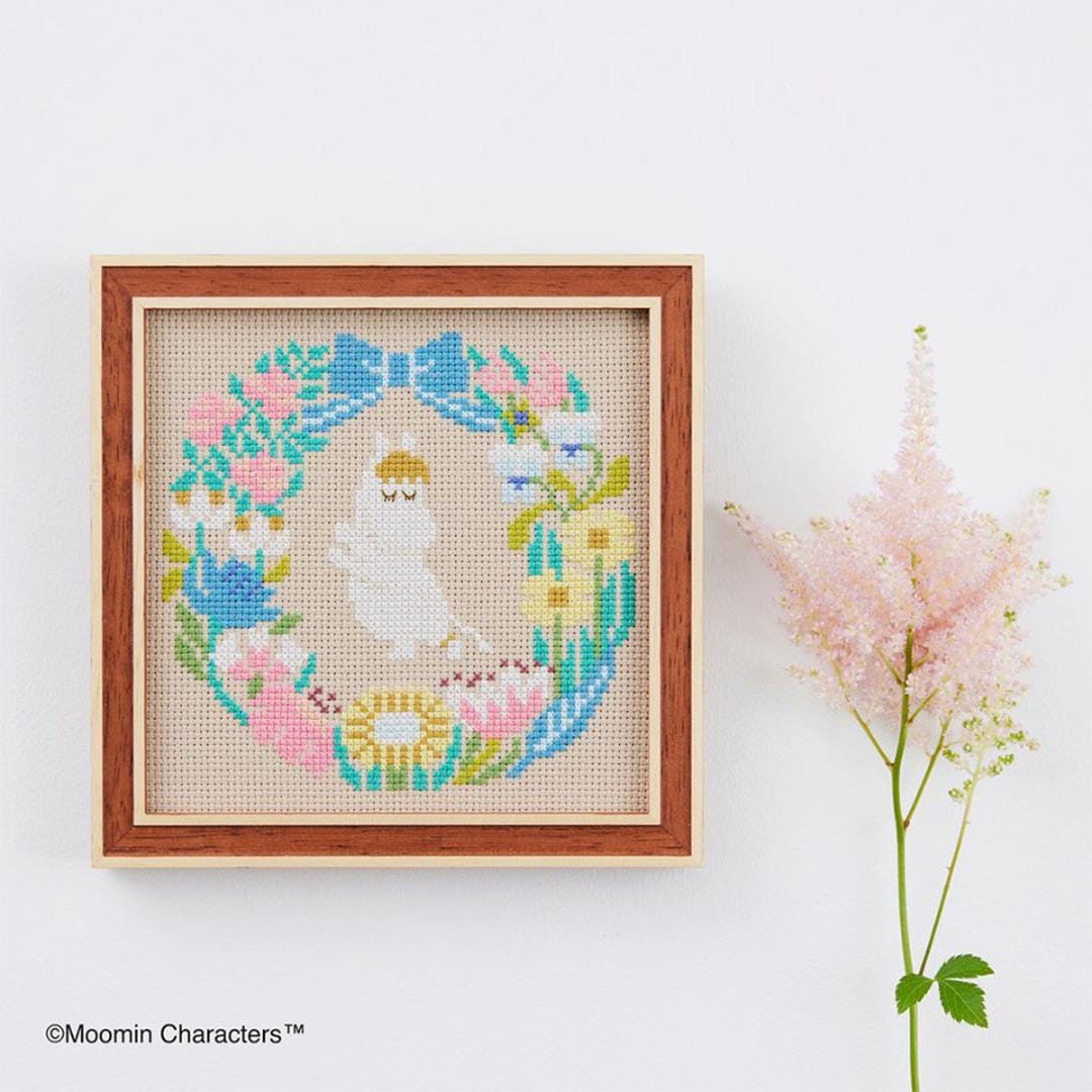 a cross-stitch design of a white cat surrounded by a floral wreath, displayed in a wooden frame on a white wall. Next to the frame is a pink flower arrangement.