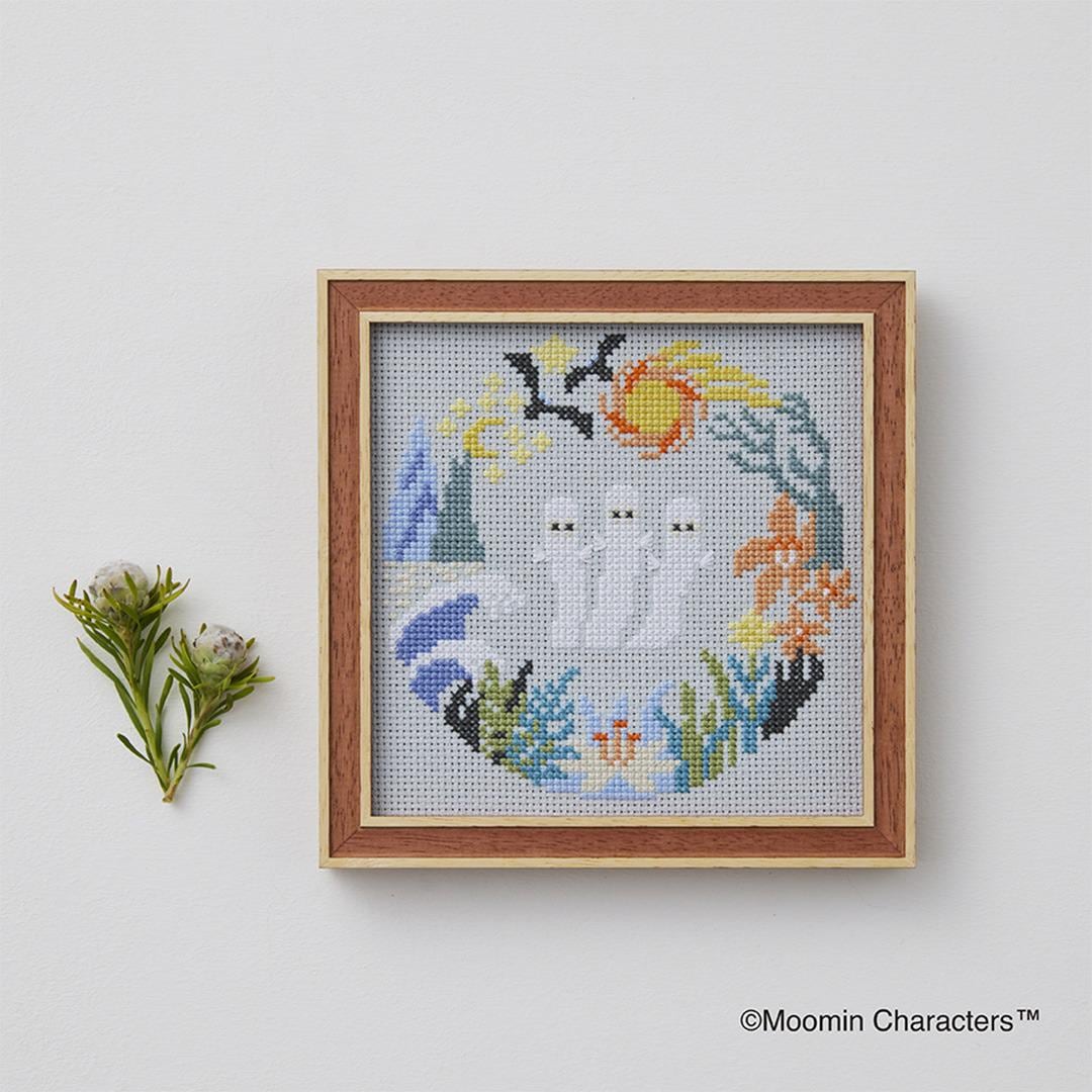 a cross-stitch design with a wreath-like border, depicting three ghostly figures surrounded by various floral and natural elements. The design is framed and displayed on a white wall, with a small plant or flower arrangement placed next to it.