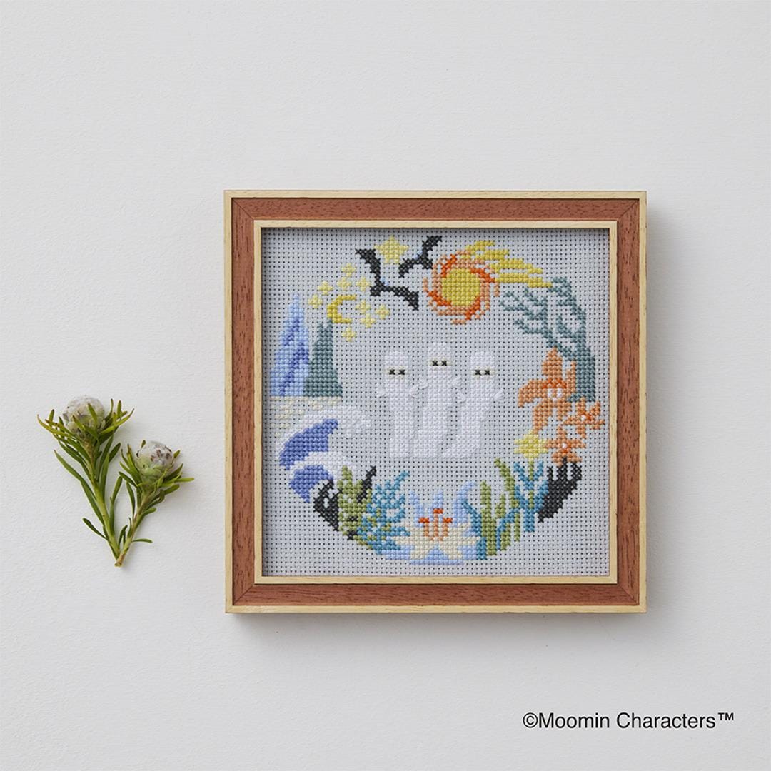 a cross-stitch design with a wreath-like border, depicting three ghostly figures surrounded by various floral and natural elements. The design is framed and displayed on a white wall, with a small plant or flower arrangement placed next to it.