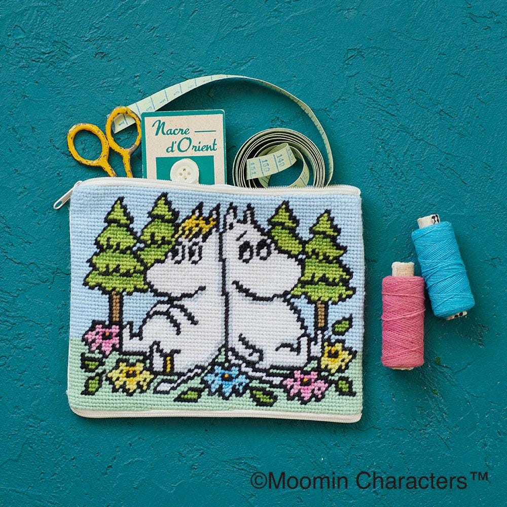 A cross-stitch pouch featuring two white characters, likely Moomins, sitting on a forest scene with trees and flowers. The pouch is placed on a teal background, and there are sewing supplies such as a spool of thread, scissors, and a measuring tape visible in the image.