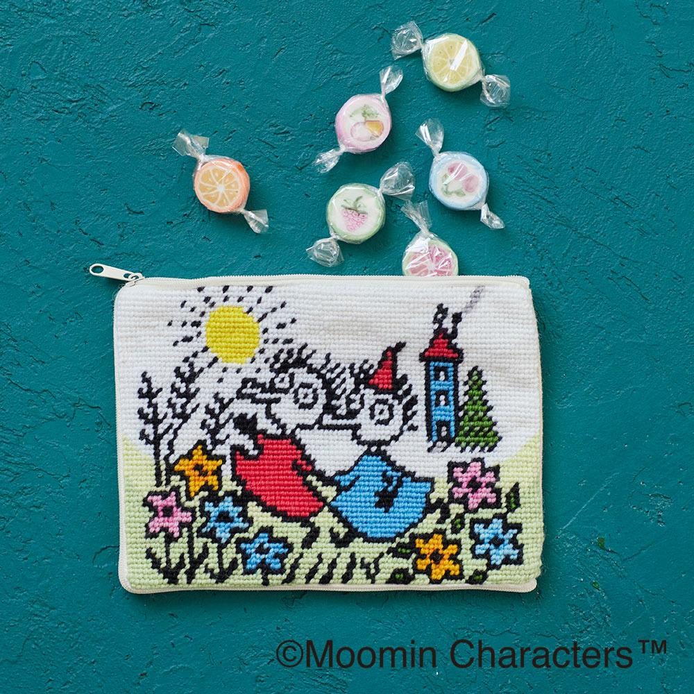 A cross-stitch pouch featuring a whimsical scene of a character surrounded by flowers and a castle, with a few colorful lollipops scattered around it.