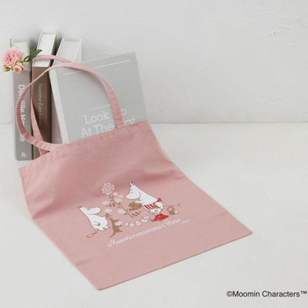 A pink tote bag with a cartoon illustration of a group of animals, including a cat, dog, and rabbit, along with the text "Moominmoomin's Flower". The bag is placed next to a stack of books, and there is a pink flower on the left side of the image.