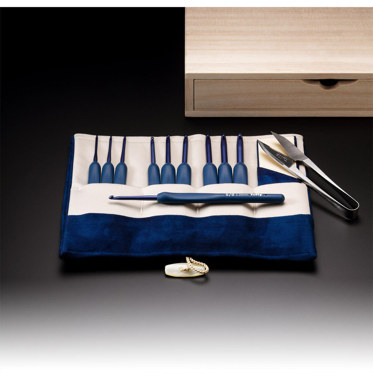 A crochet set consisting of a crochet hook and a pair of scissors is displayed on a blue and white cloth, with a wooden box and a pair of scissors in the background.