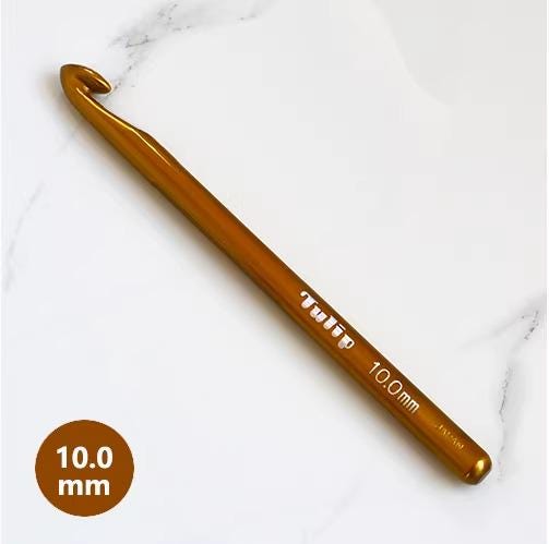 A gold crochet hook with the text "Tulip 10.0mm" printed on it, resting on a white marble surface.