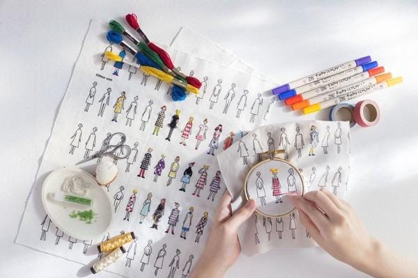 a person's hands holding a embroidery hoop with a design of various stylized human figures, surrounded by sewing supplies and materials.