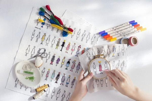 a person's hands holding a embroidery hoop with a design of various stylized human figures, surrounded by sewing supplies and materials.