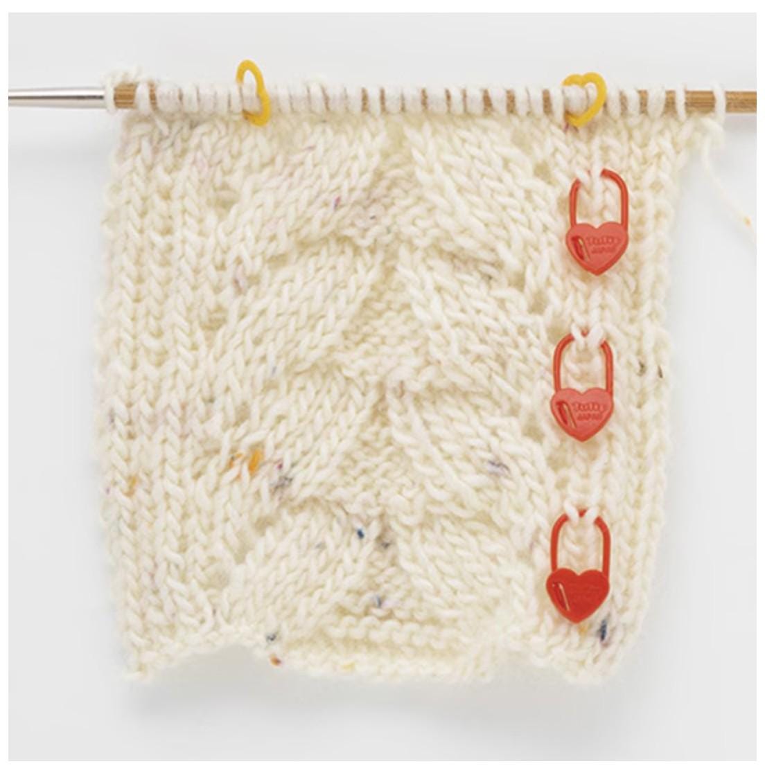 A close-up view of a knitted fabric with red heart-shaped tags attached to it, hanging from a knitting needle.