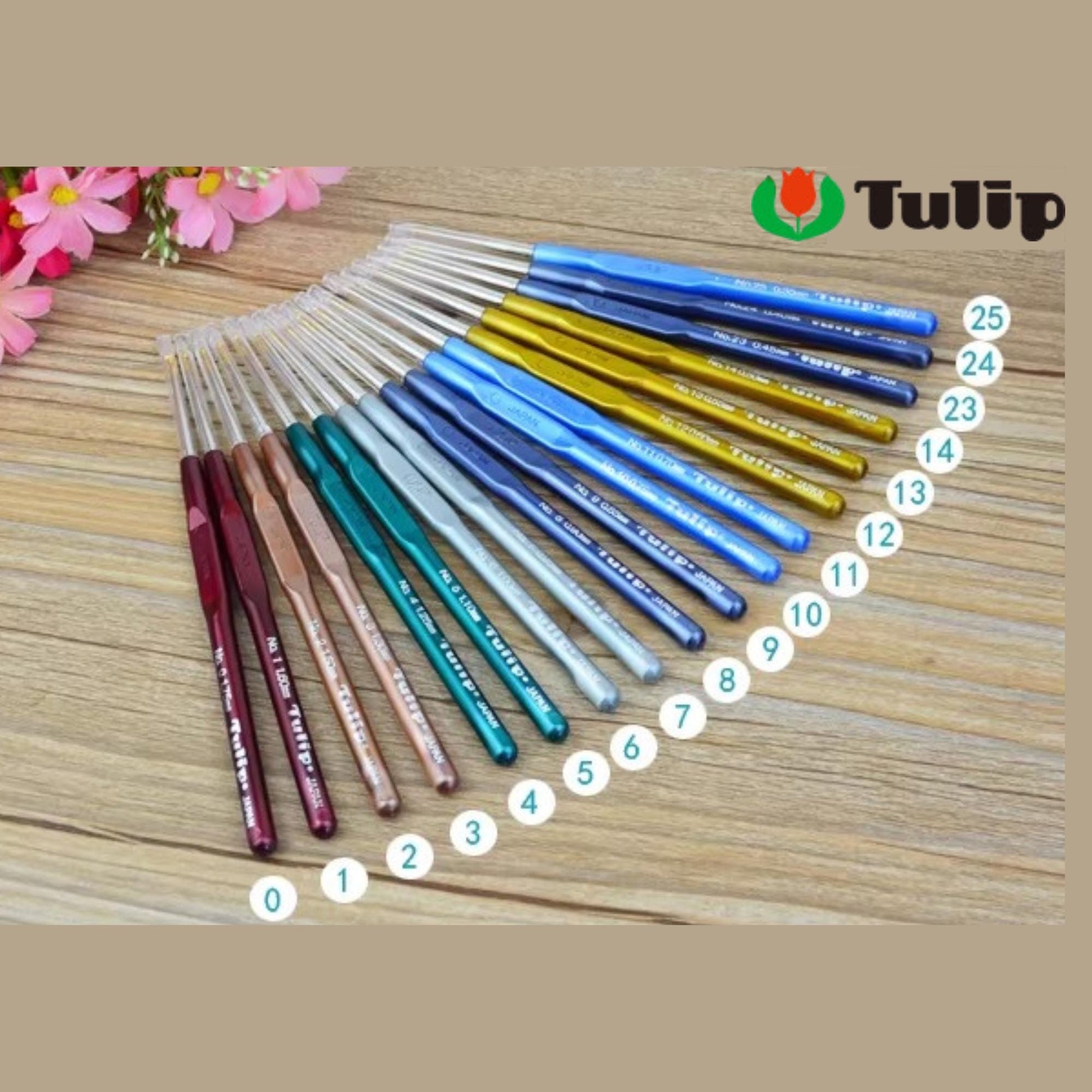 A collection of colorful crochet hooks arranged on a wooden surface, with pink flowers in the background.