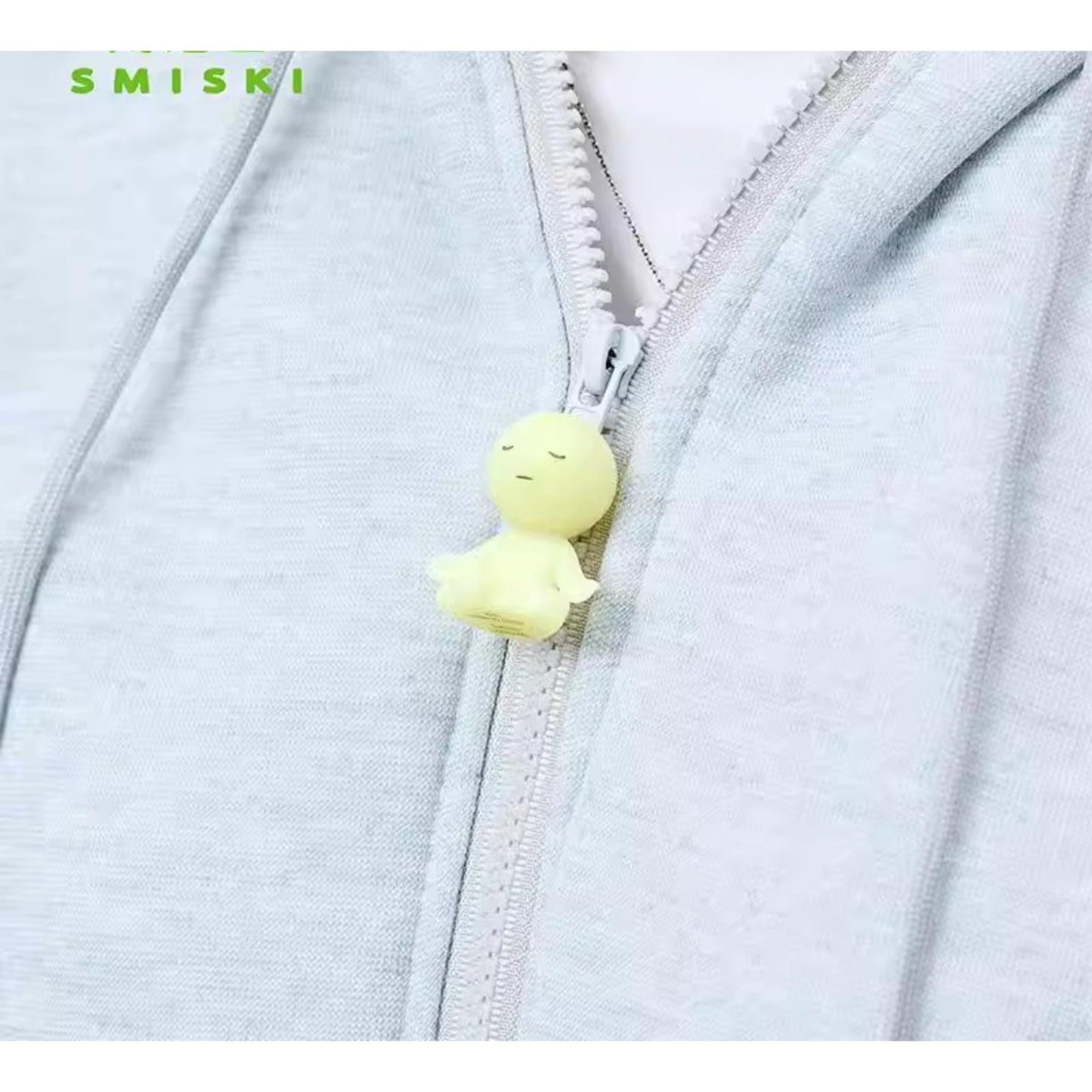A small yellow figurine is attached to a zipper on a gray hoodie.