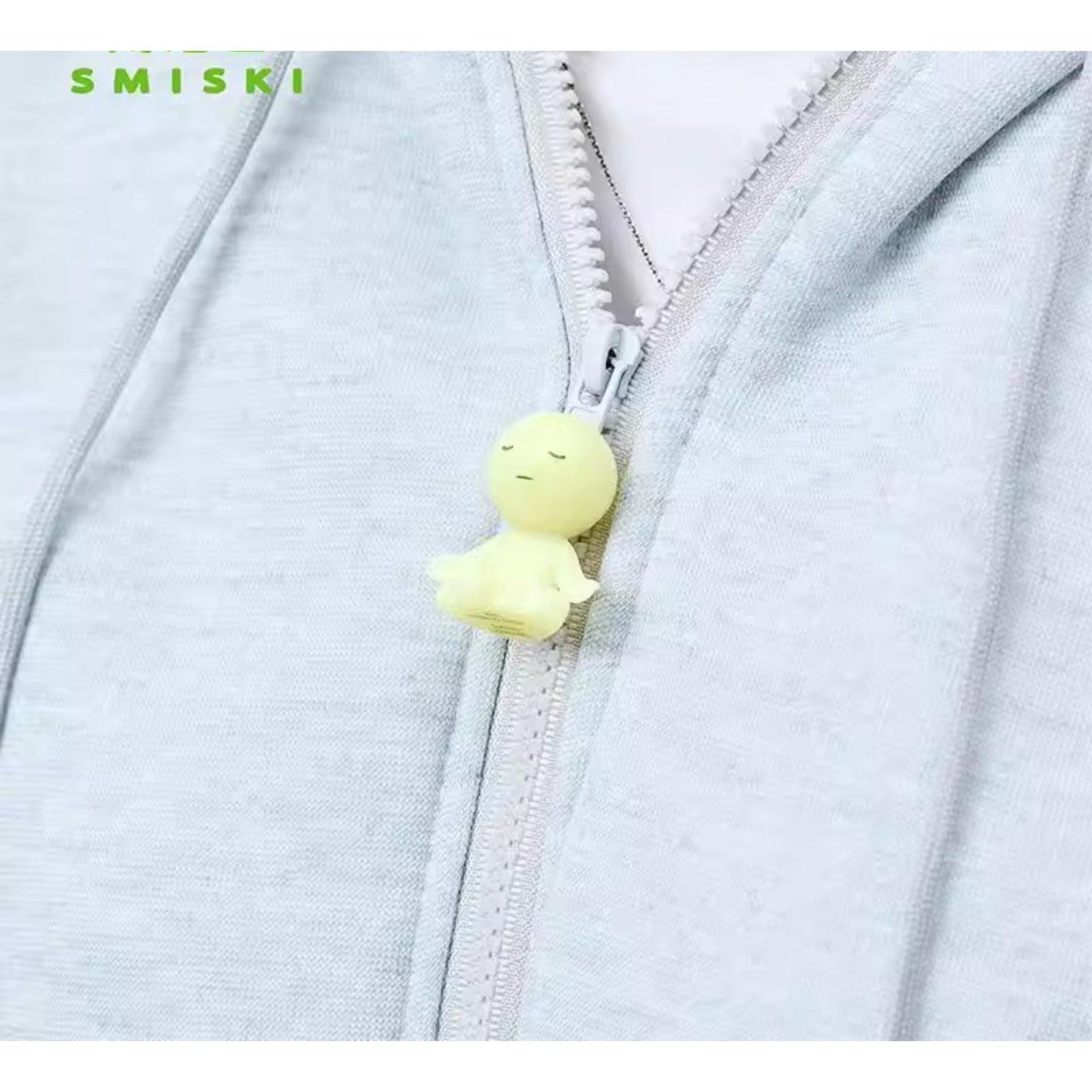 A small yellow figurine is attached to a zipper on a gray hoodie.