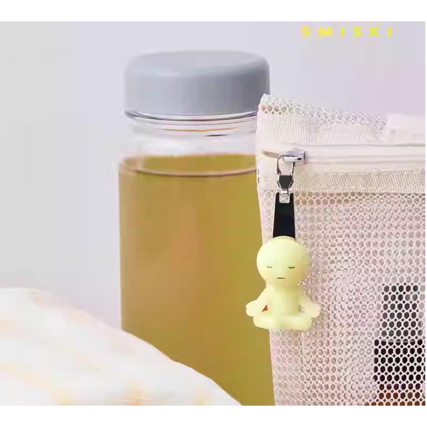 a small yellow figurine hanging from a mesh bag, with a glass bottle and a white mesh bag visible in the background.