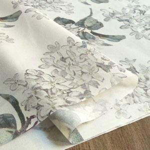 a close-up view of a fabric with a floral pattern, featuring white flowers and leaves. The fabric appears to be folded or rolled, with the edges slightly curled. The background is blurred, drawing focus to the fabric with the floral pattern.