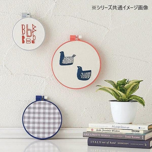 a white wall with three embroidery hoops hanging on it. The first hoop has a red and white design, the second hoop has a blue and white design, and the third hoop has a checkered pattern. A potted plant is placed on top of a stack of books, and there are some books visible in the background.