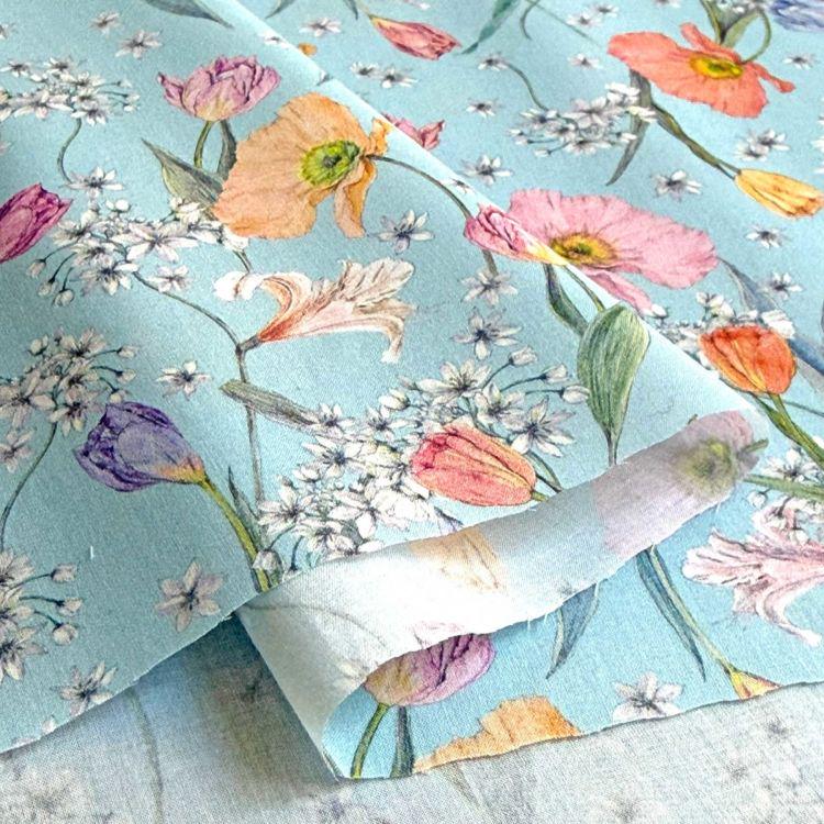 a close-up view of a fabric with a vibrant floral pattern. The fabric is predominantly blue, with various flowers such as tulips, lilies, and daisies in shades of pink, orange, and white. The flowers are arranged in a way that creates a visually appealing and colorful design.