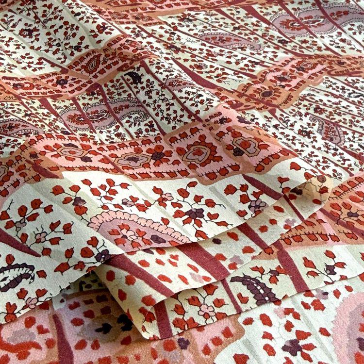 The image is a close-up of a fabric with a repeating pattern of red and white floral designs. The pattern is intricate and detailed, with various shapes and sizes of flowers and leaves. The fabric appears to be folded or rolled, revealing the intricate design underneath.