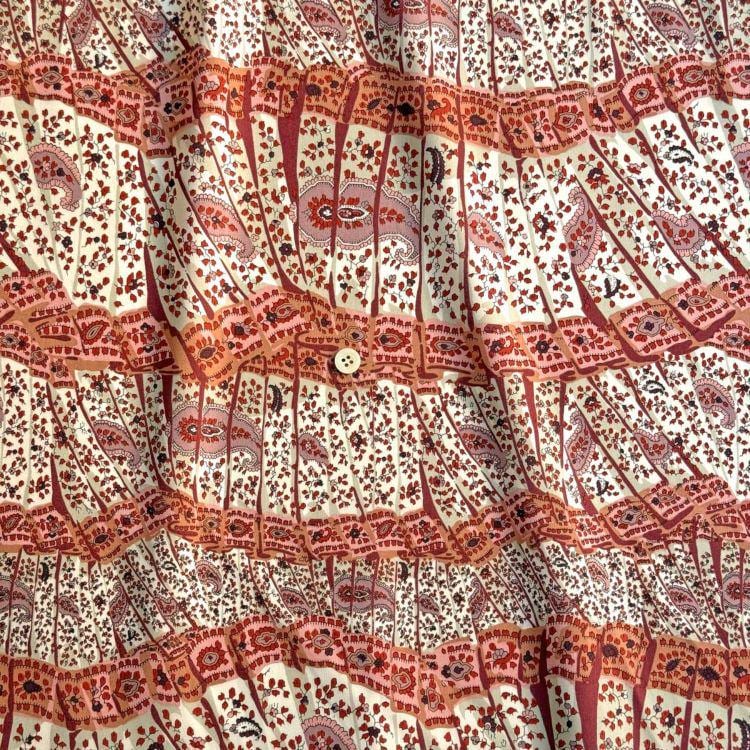 a close-up view of a fabric with a repeating pattern of red and white paisley designs. The fabric appears to be made of a soft, flowy material, and there is a button visible in the center of the pattern.