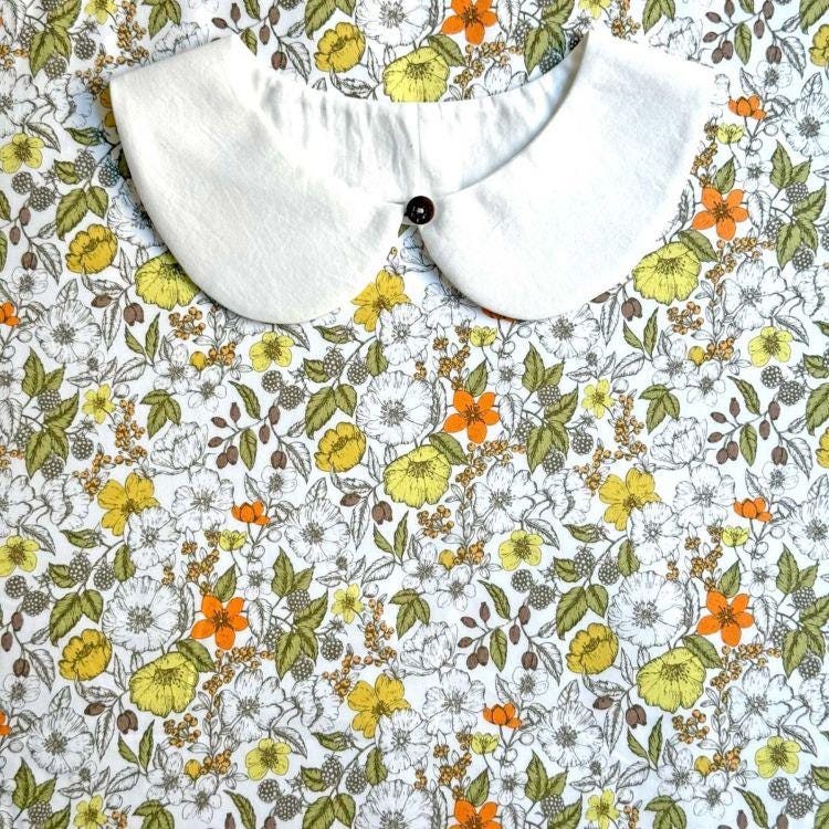 a white collar with a black button, placed on a fabric with a floral pattern of yellow, orange, and white flowers.