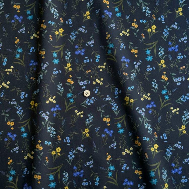 a close-up view of a fabric with a floral pattern in shades of blue, yellow, and green. The fabric appears to be dark blue, and the pattern consists of small flowers and leaves in various colors. There is a button visible in the center of the fabric.