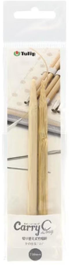 A package containing a pair of wooden knitting needles, with a blurred background that appears to be a sewing or crafting workspace.