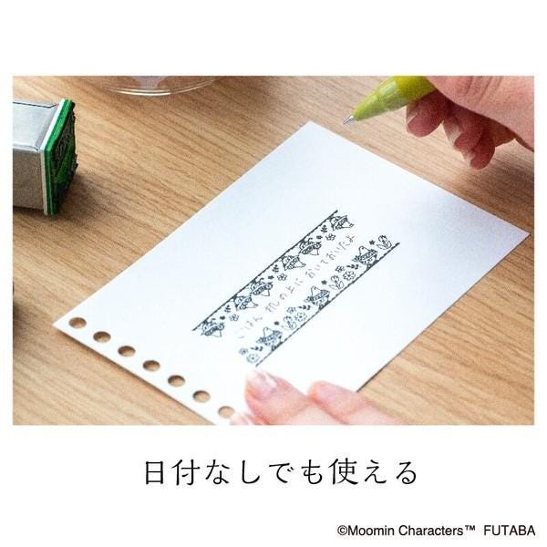 A hand is holding a green pen and writing on a piece of paper with Japanese characters, which appears to be a note or a message.