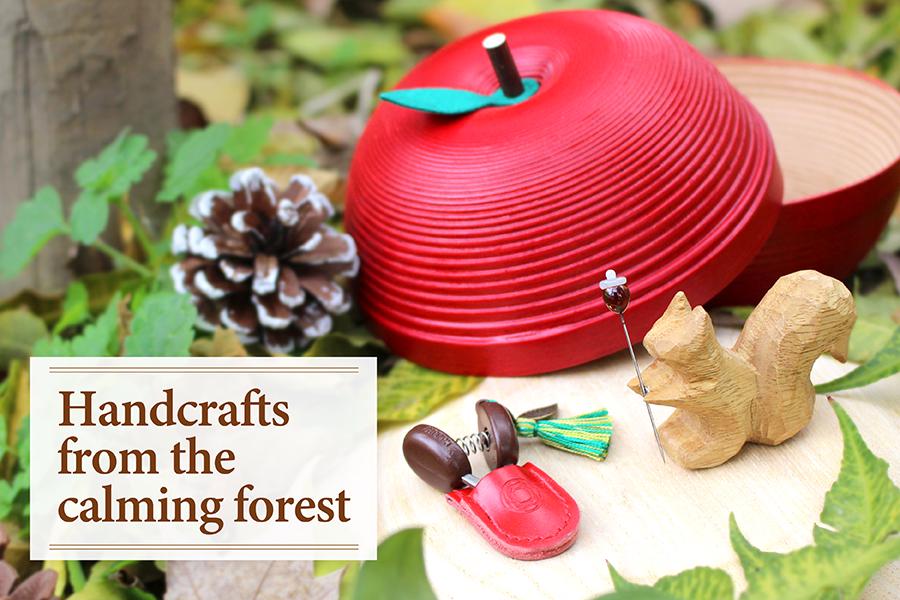 a red apple-shaped container, a pine cone, a wooden squirrel figurine, and a red keychain, all placed on a wooden surface surrounded by leaves and branches, with a sign that reads "Handcrafts from the calming forest".