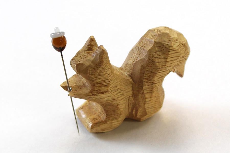 A wooden squirrel figurine is shown, with a small acorn attached to its tail using a pin.