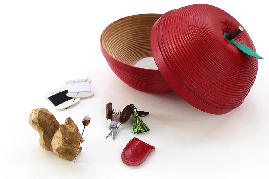 a red apple-shaped container with a lid, along with various craft supplies and tools, such as scissors, a tassel, and a small wooden sculpture.