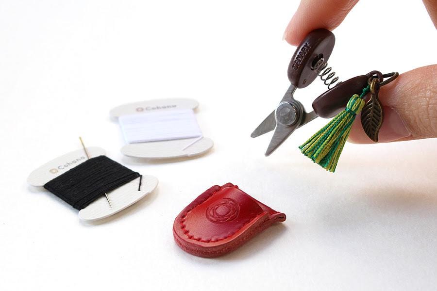 A hand holding a pair of scissors with a tassel attached, surrounded by sewing supplies such as a spool of thread, a needle, and a small red leather pouch.