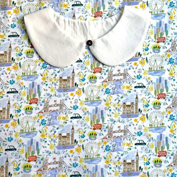 a white collar with a button, placed on a fabric with a colorful, whimsical pattern of various landmarks and city scenes, including ferris wheels, bridges, and buildings.