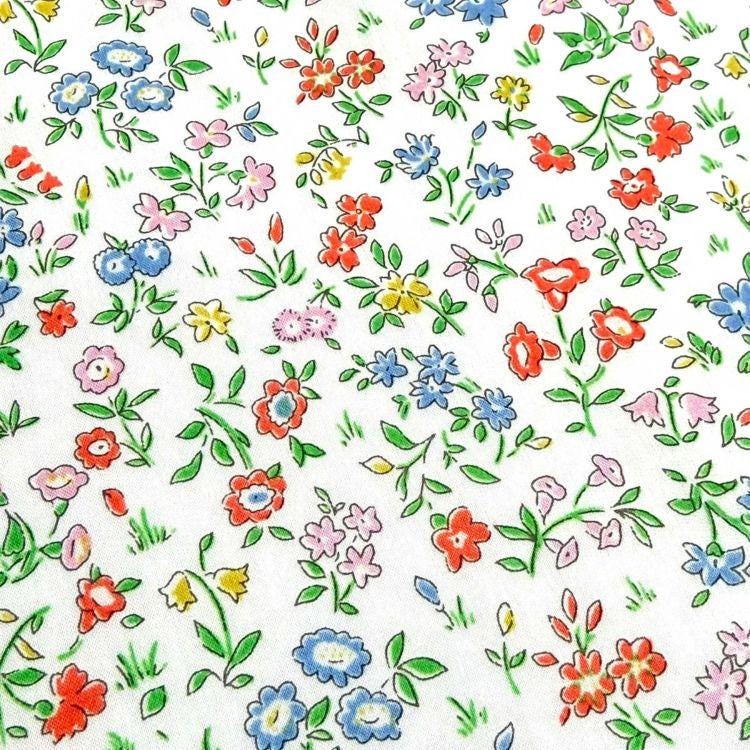 The image is a close-up of a white fabric with a vibrant floral pattern. The pattern consists of various types of flowers, including red, pink, blue, and yellow flowers, as well as green leaves. The flowers are scattered across the fabric in a random and overlapping manner, creating a visually appealing and lively design.