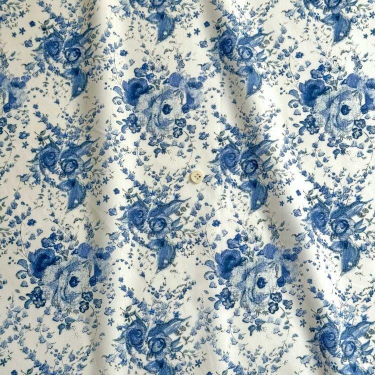 a close-up view of a fabric with a repeating floral pattern in shades of blue and white. The pattern consists of various flowers and leaves, creating a visually appealing and intricate design. The fabric appears to be made of a soft, textured material, suitable for clothing or home decor.