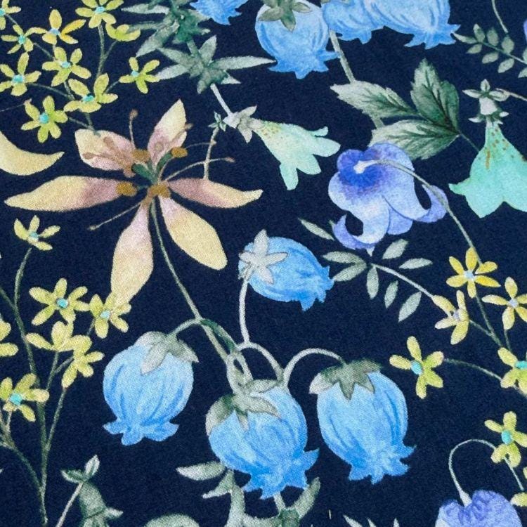 a close-up view of a dark blue fabric adorned with a vibrant floral pattern. The pattern includes various types of flowers, such as bluebells, daisies, and lilies, along with leaves and stems. The colors of the flowers and leaves contrast beautifully against the dark blue background, creating a visually striking and intricate design.