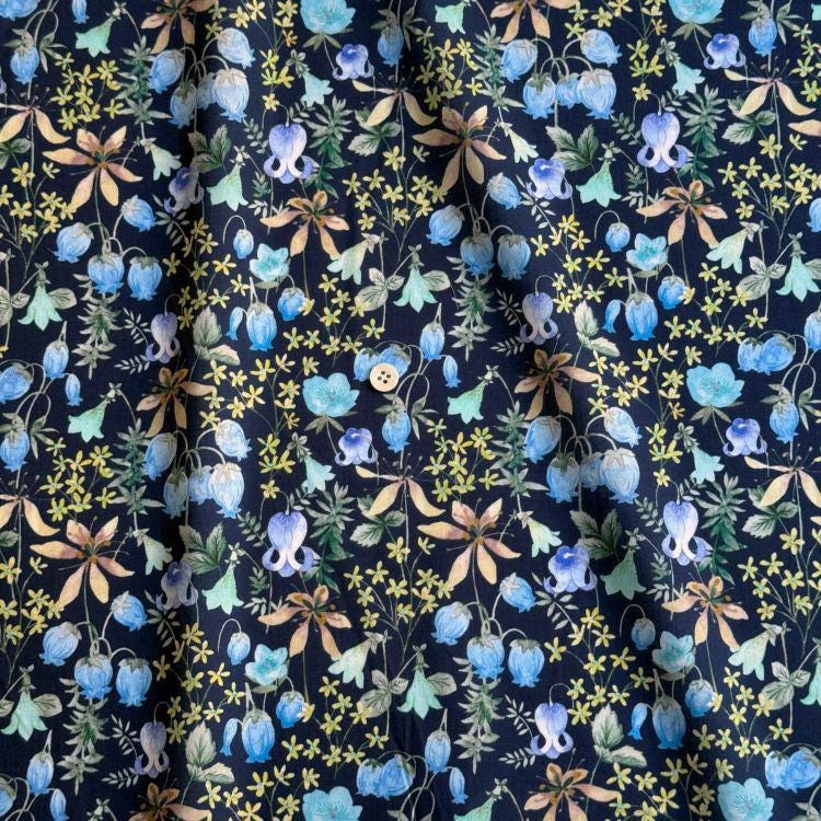 a close-up view of a fabric with a floral pattern in shades of blue, yellow, and green. The pattern consists of various flowers and leaves, including bluebells, daisies, and other types of flowers, all set against a dark background. The fabric appears to be a dark-colored material, possibly cotton or linen, with a subtle texture that adds depth to the overall design.