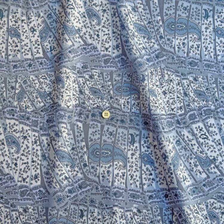 a close-up view of a fabric with a repeating pattern of blue and white paisley designs. The fabric appears to be made of a soft, textured material, and there is a small yellow button visible in the center of the pattern.
