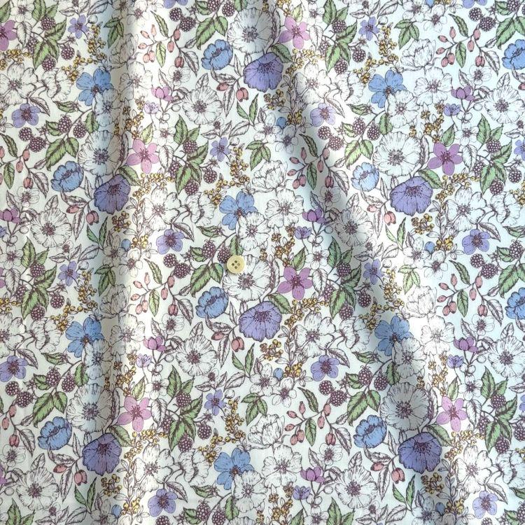 a close-up view of a fabric with a floral pattern. The pattern consists of various flowers and leaves in different colors, including white, purple, and blue. The fabric appears to be made of a soft, white material, and the pattern is intricate and detailed.