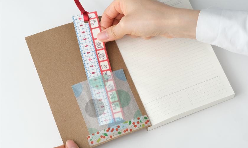 A hand is holding a red ruler and a brown notebook, with the ruler partially inserted into the notebook.