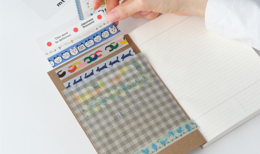 A hand is holding a pack of colorful washi tape, with the tape peeling off a notebook.