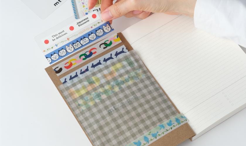 A hand is holding a pack of colorful washi tape, with the tape peeling off a notebook.