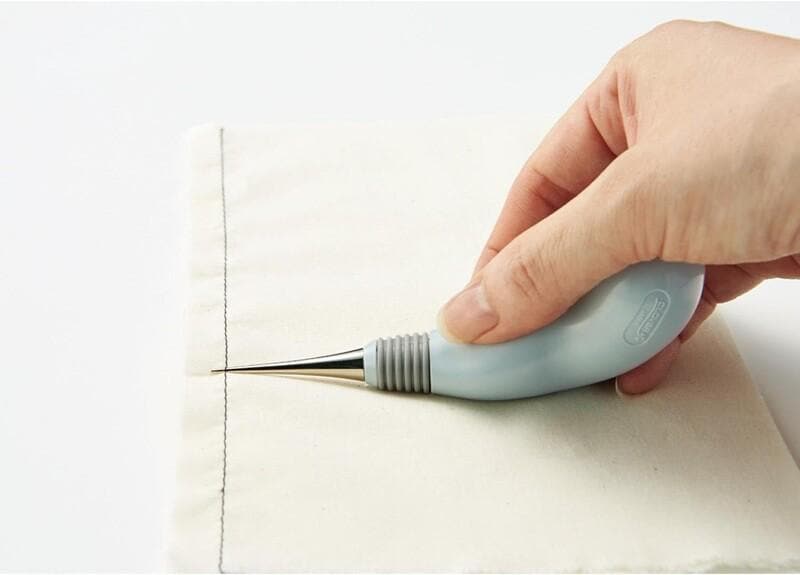 A hand is using a blue and silver tool to carefully cut a white fabric, likely for a sewing or crafting project.